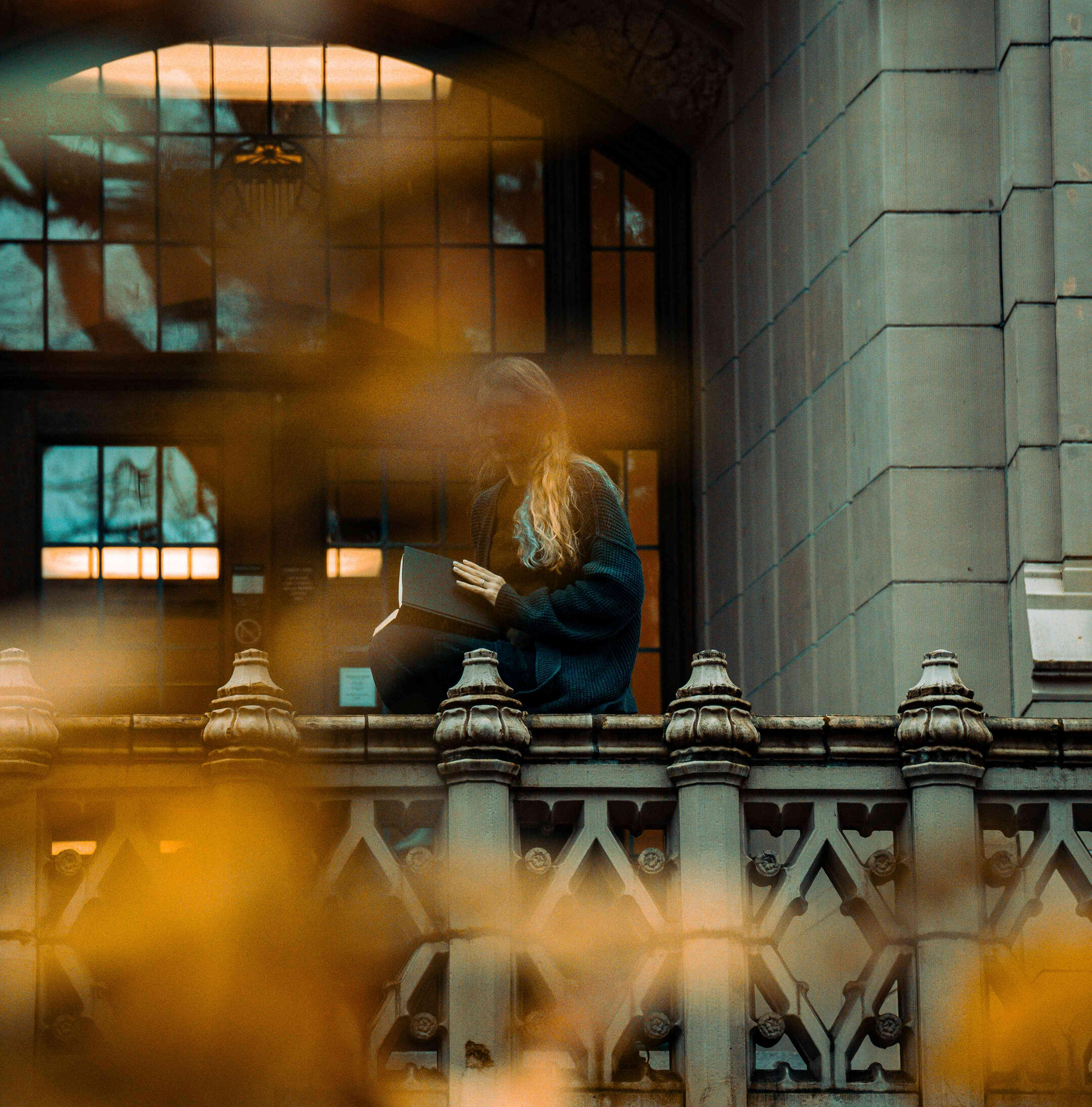 A woman reads along a balcony. She is obscured through yellow and orange leaves in the distance. Photo credit: https://www.instagram.com/hello_meagain/