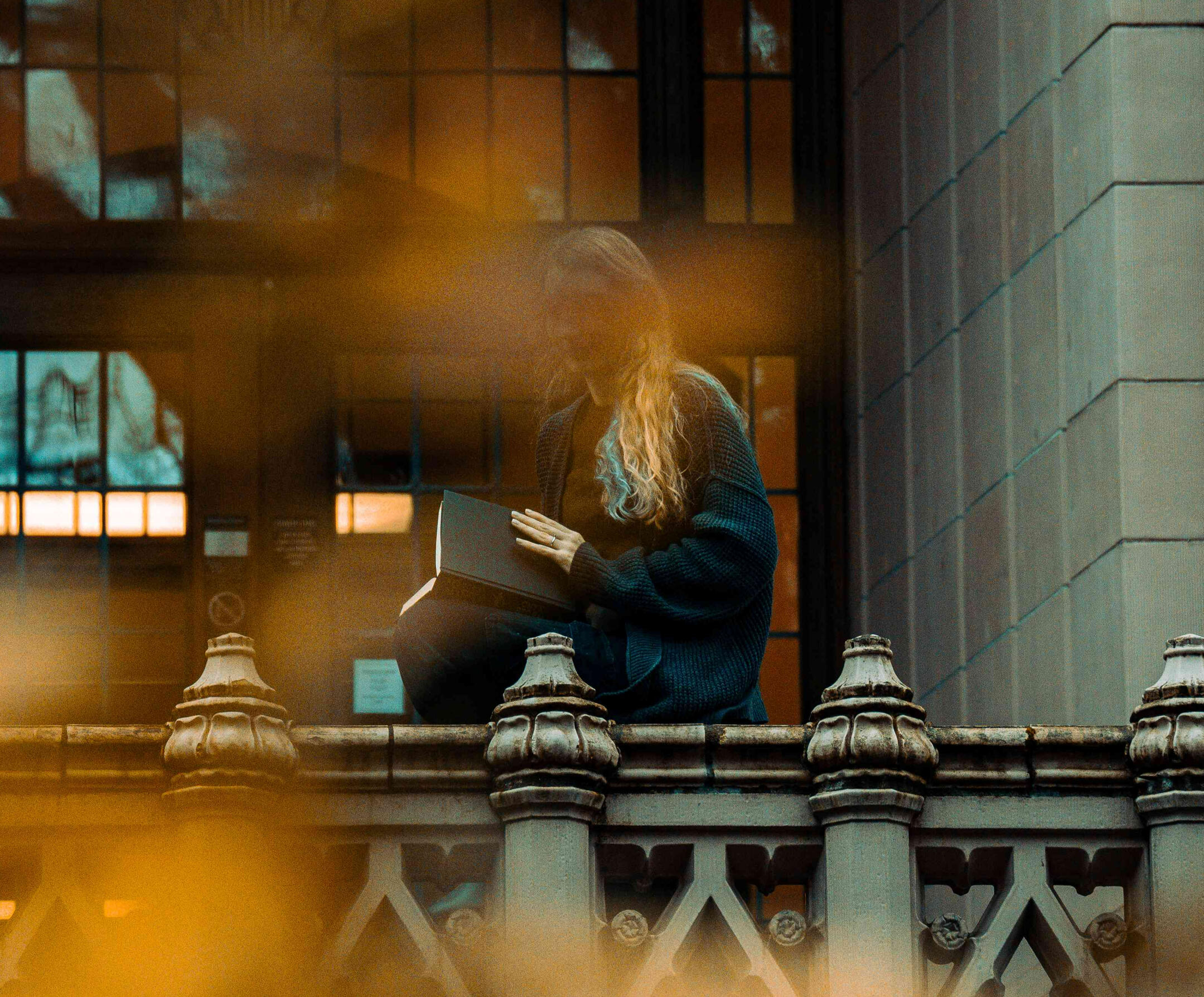 A woman reads along a balcony. She is obscured through yellow and orange leaves in the distance. Photo credit: https://www.instagram.com/hello_meagain/